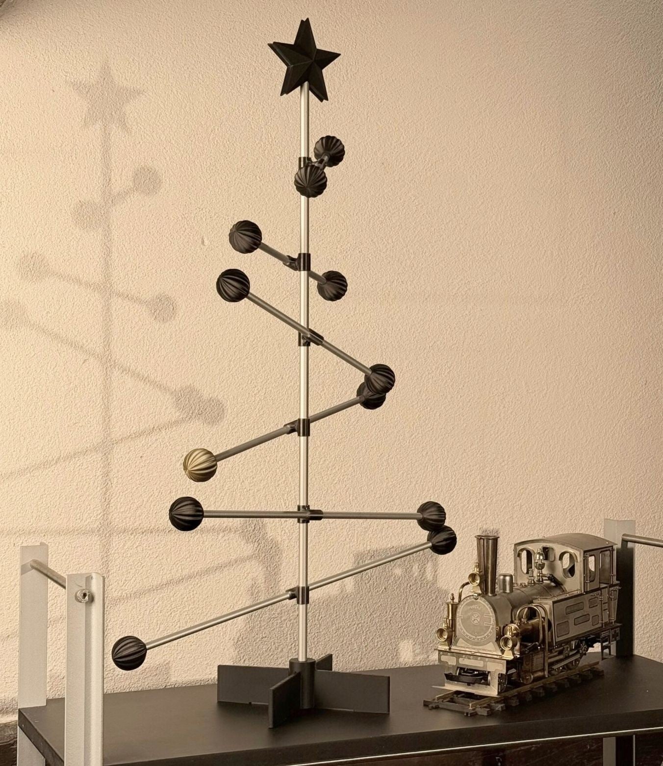 Decorative metal Christmas tree with a black star on top, placed on a surface against a neutral wall. A steam engine train next to it.