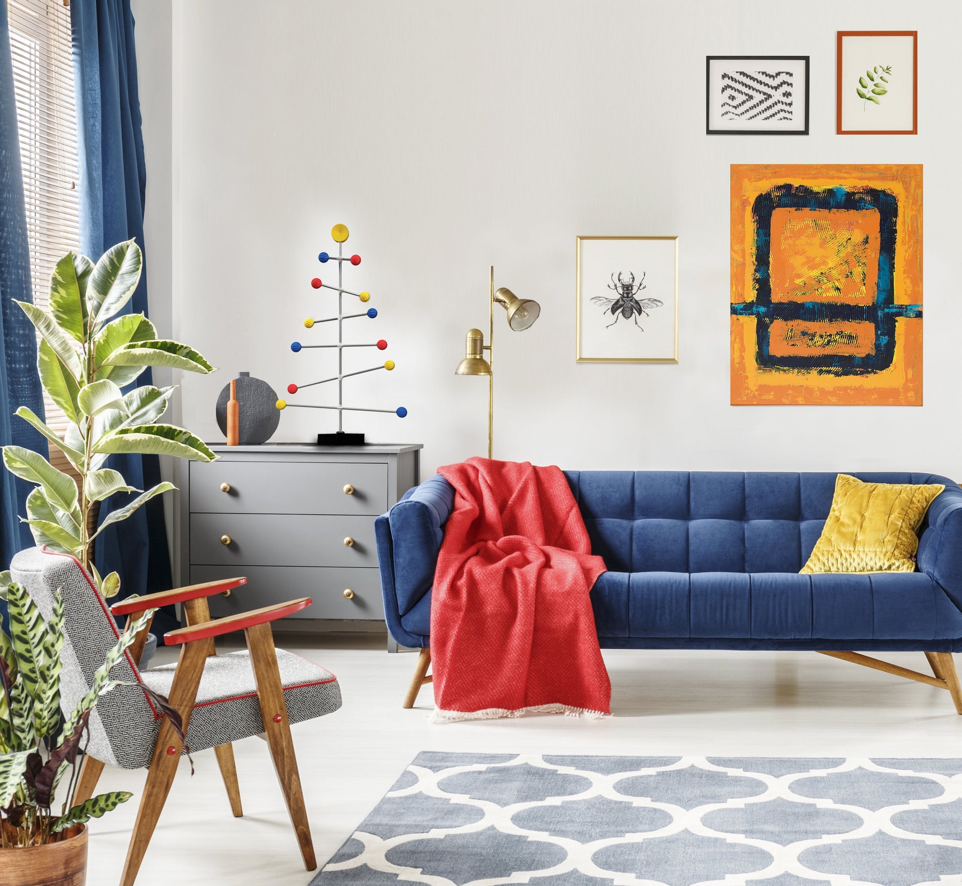 Wohnzimmer mit blauem Sofa, roter Decke und bunten Kunstwerken an der Wand. Und ein Weihnachtsbaumständer in Bauhaus-Farben auf dem Schrank.