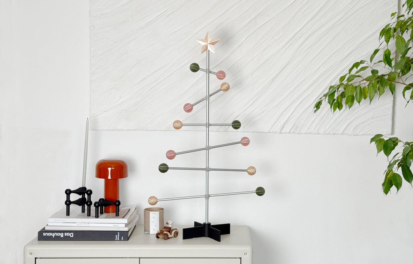 Modern decorative easter /Christmas tree with pastel colored balls on a white surface next to books and a plant.
