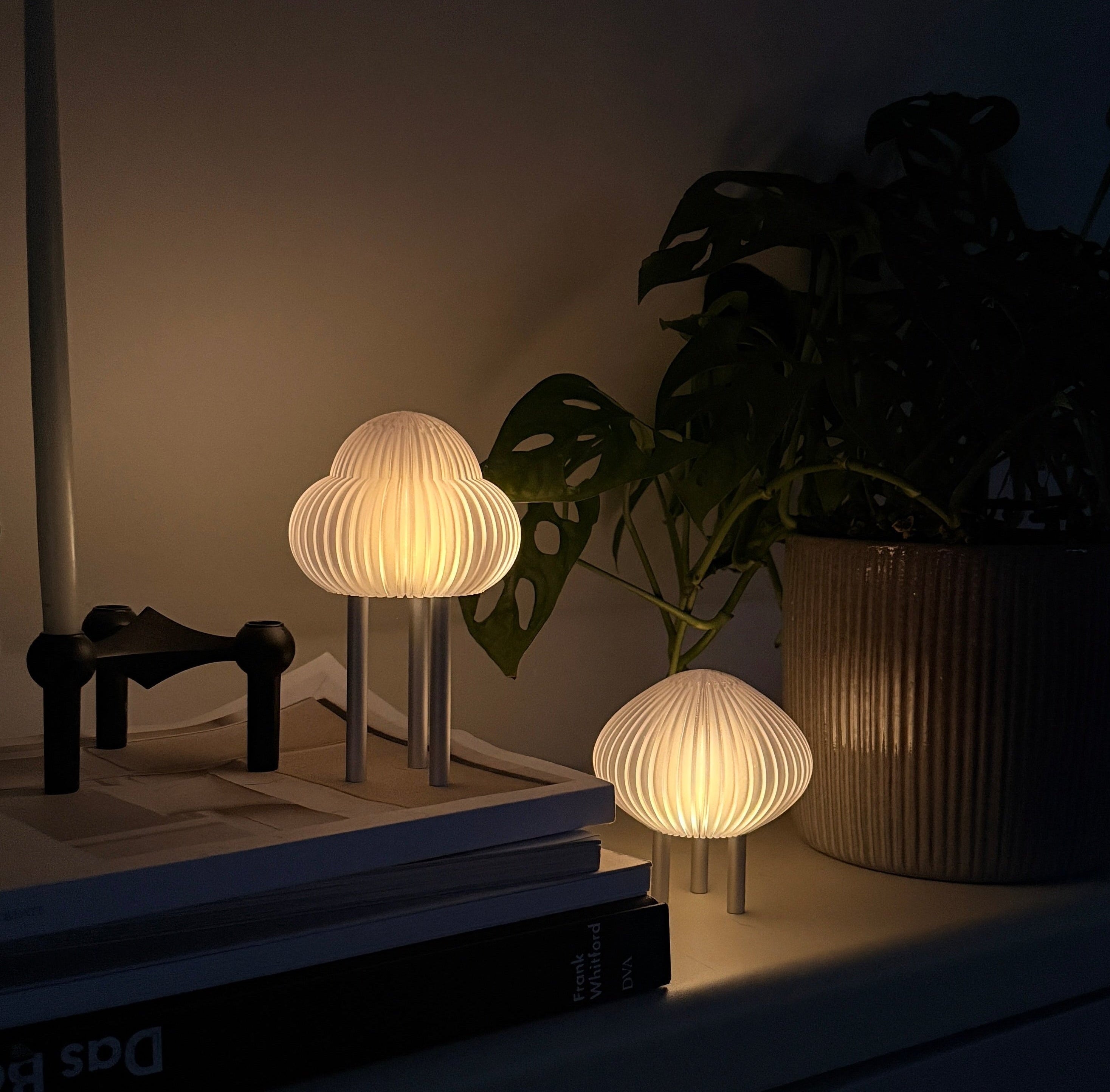 Two small mushroom-shaped table lamps with pleated white shades and slim silver bases, placed on a dark shelf beside a potted plant in a dimly lit room.
