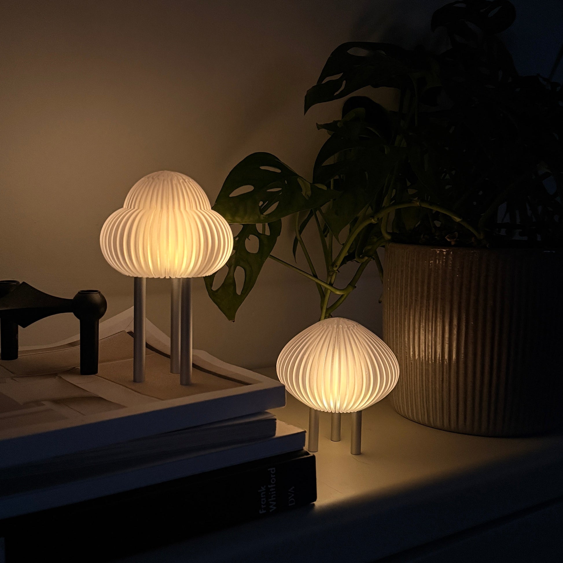 Two modern mushroom-shaped table lamps with pleated white shades and silver metal bases, shown lit on a shelf next to a potted plant in a dark room.