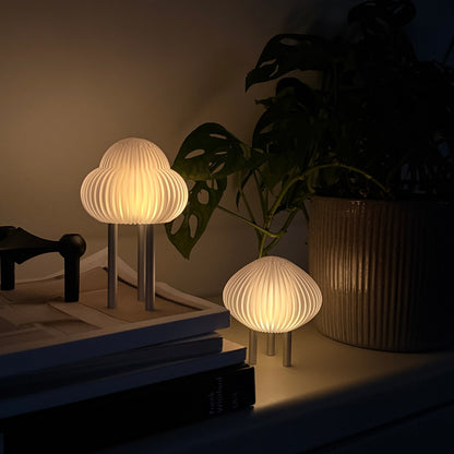 Two modern mushroom-shaped table lamps with pleated white shades and silver metal bases, shown lit on a shelf next to a potted plant in a dark room.