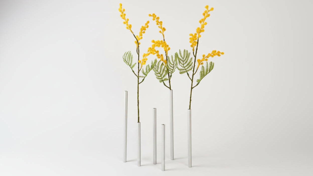 Three tall, slender silver cylindrical vases standing on a light gray surface against a neutral light gray background. Each vase holds a dried botanical stem with bright yellow flowers and green leaves.