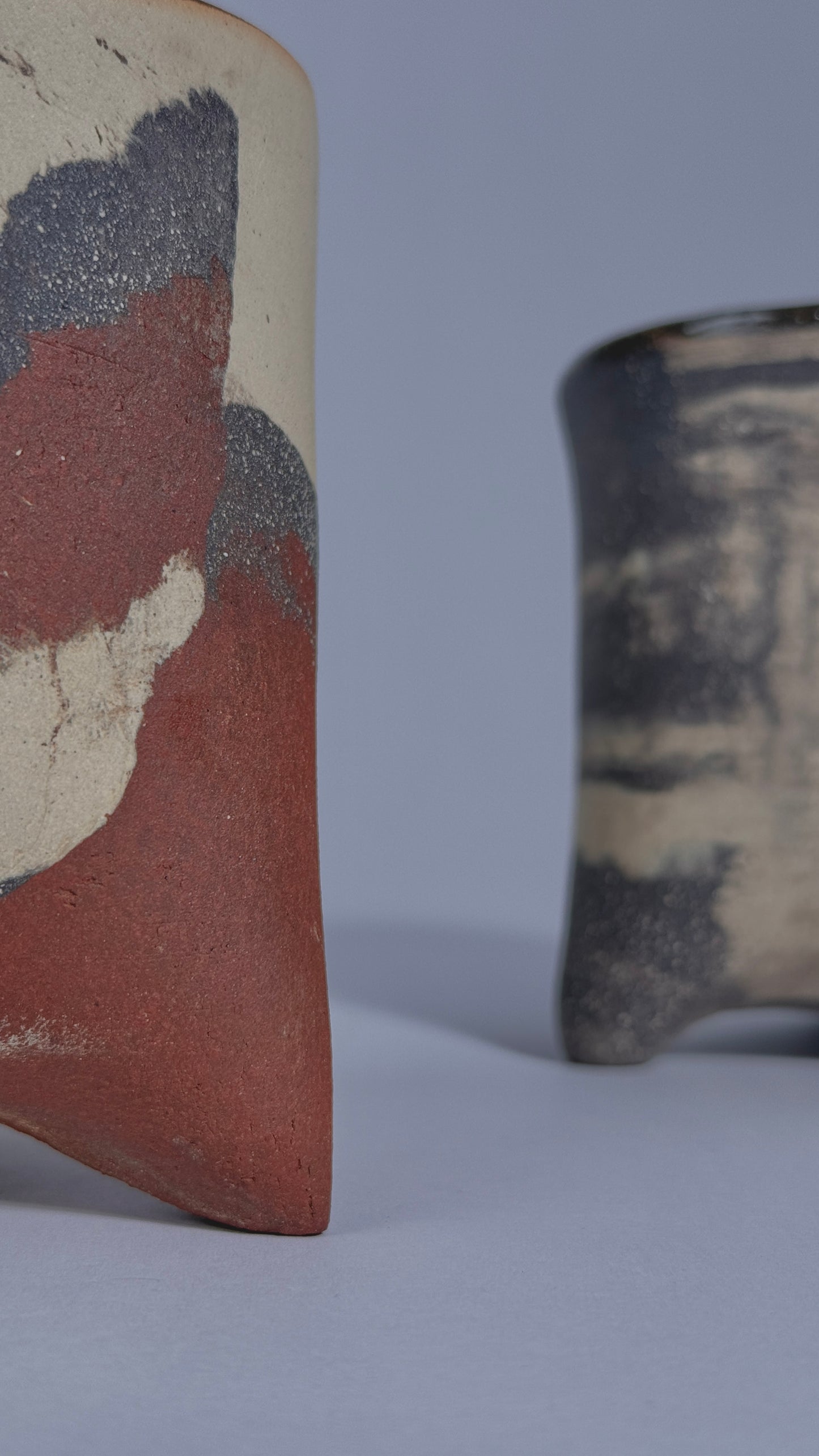 Two handmade ceramic vessels with textured, abstract glazes in earthy tones of terracotta red, charcoal gray, and cream, shown on a neutral light gray background.