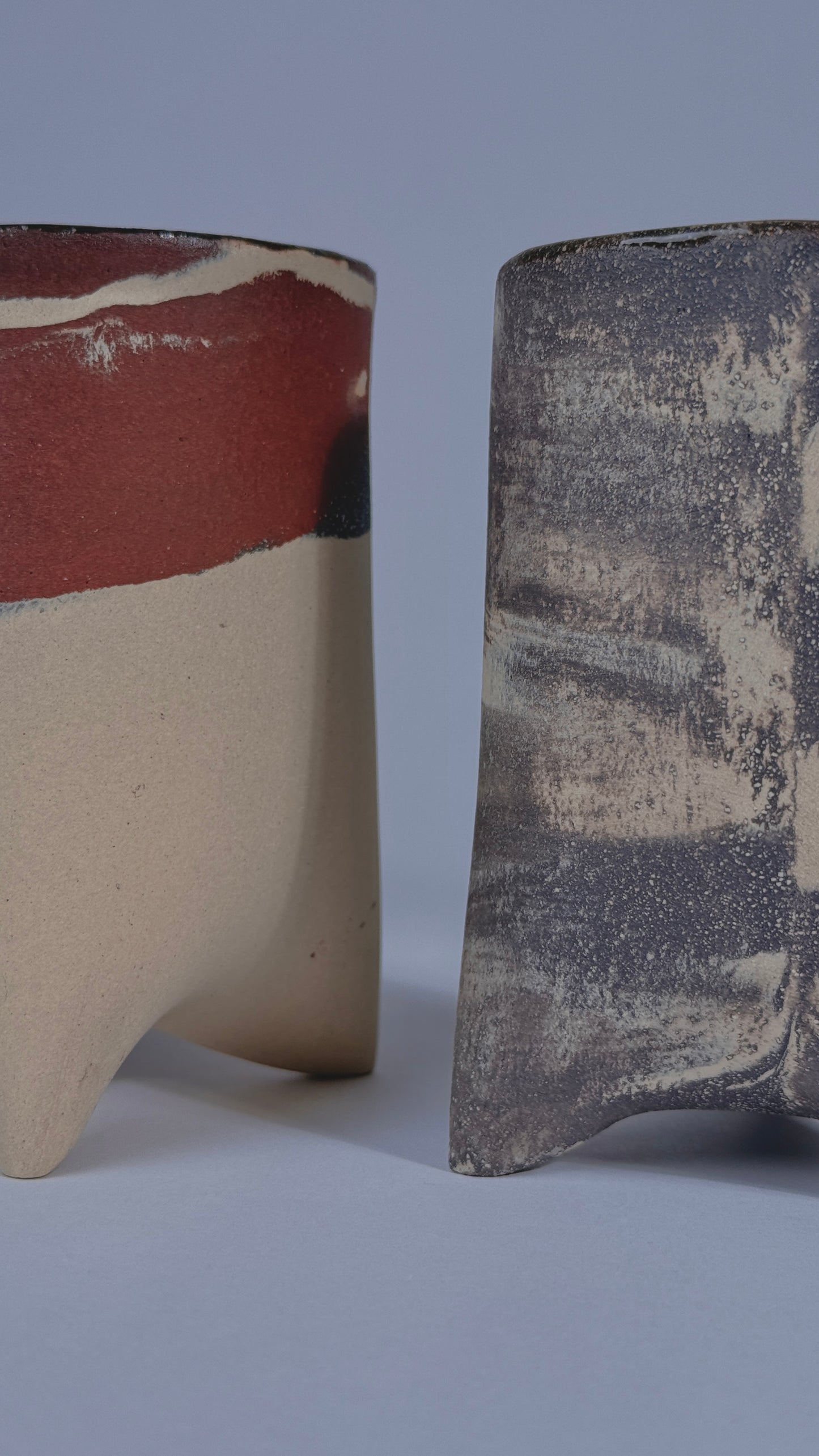 Two handmade ceramic vessels with textured, rustic glazes shown side-by-side against a neutral gray background.