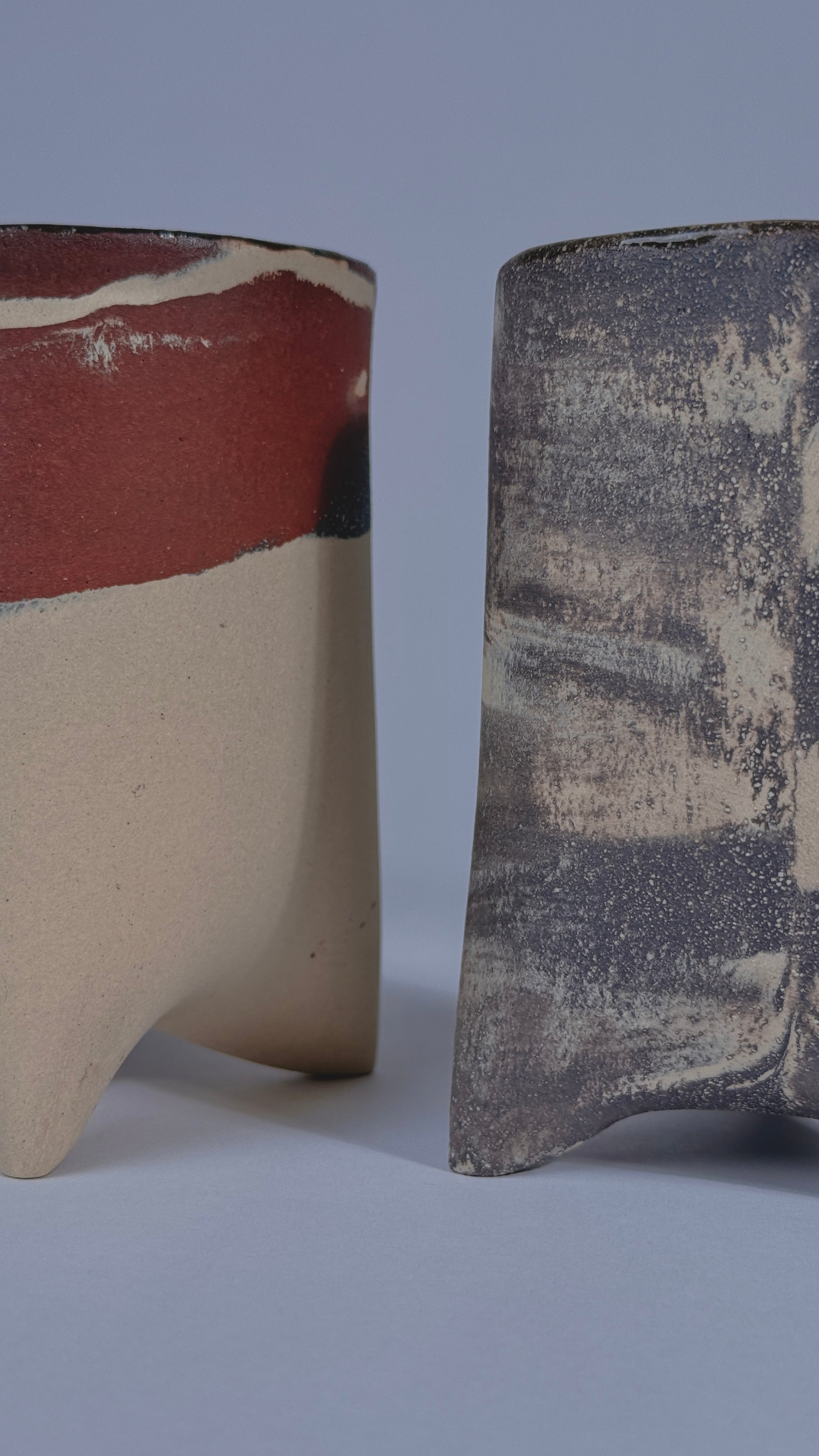 Two handmade ceramic vessels with textured, rustic glazes shown side-by-side against a neutral gray background.