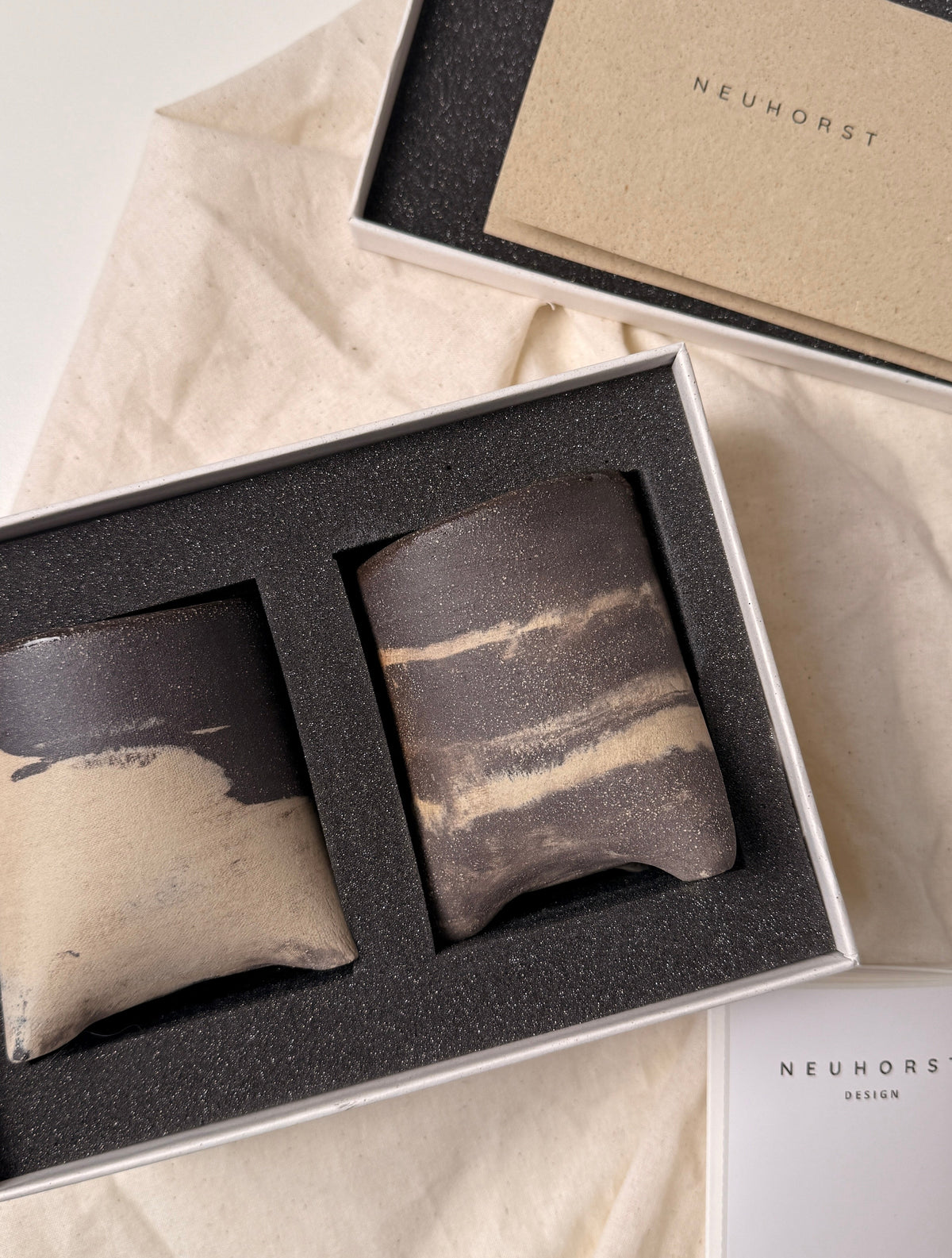 A gift set of two ceramic coasters in a white box with black foam insert, displayed on beige fabric alongside branded packaging cards reading NEUNHORST.