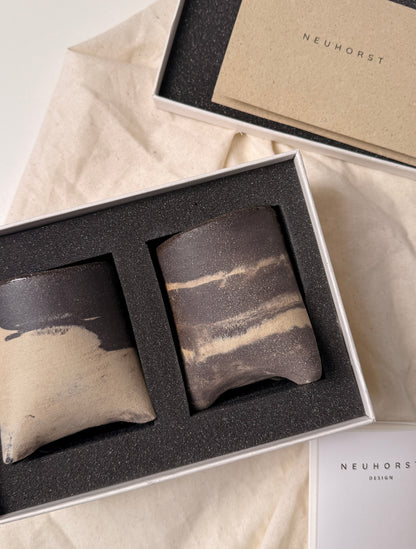A gift set of two ceramic coasters in a white box with black foam insert, displayed on beige fabric alongside branded packaging cards reading NEUNHORST.
