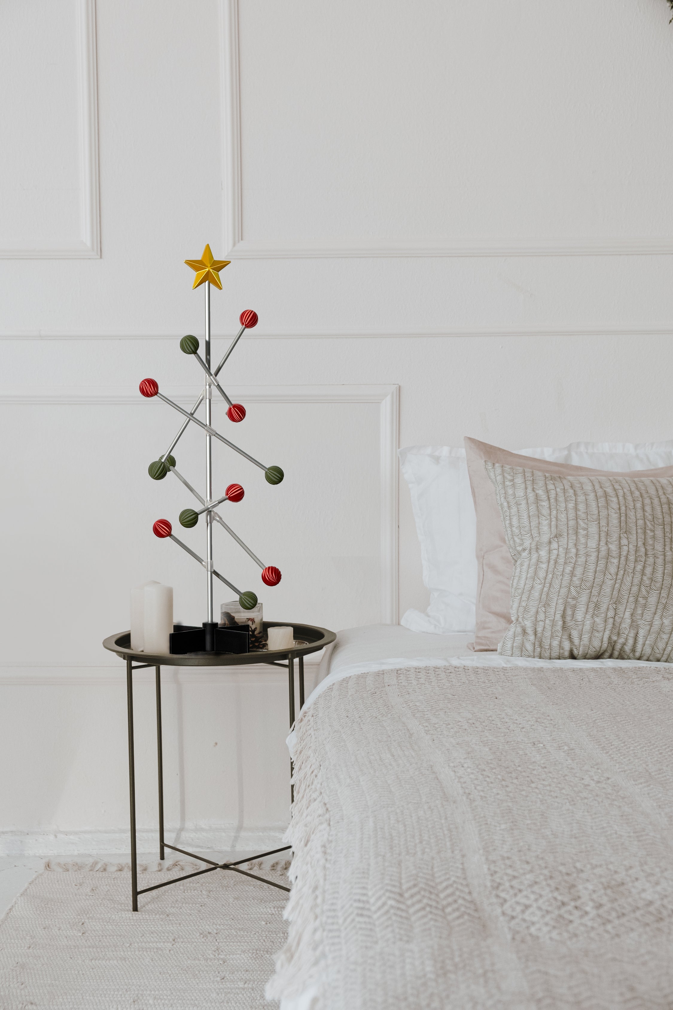 Small Christmas tree with red and green spheres and gold star decorations on a bedside table next to a bed with white bedding.