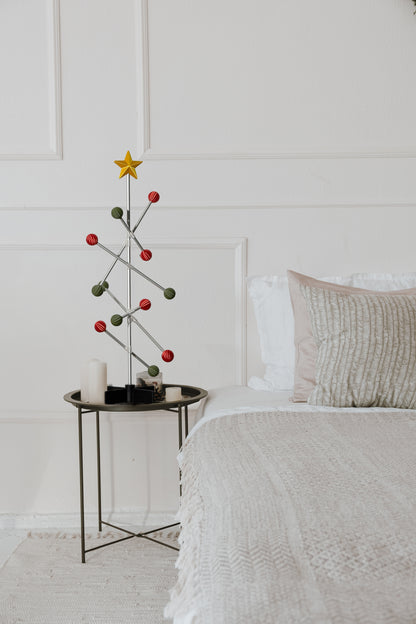 Small Christmas tree with red and green spheres and gold star decorations on a bedside table next to a bed with white bedding.